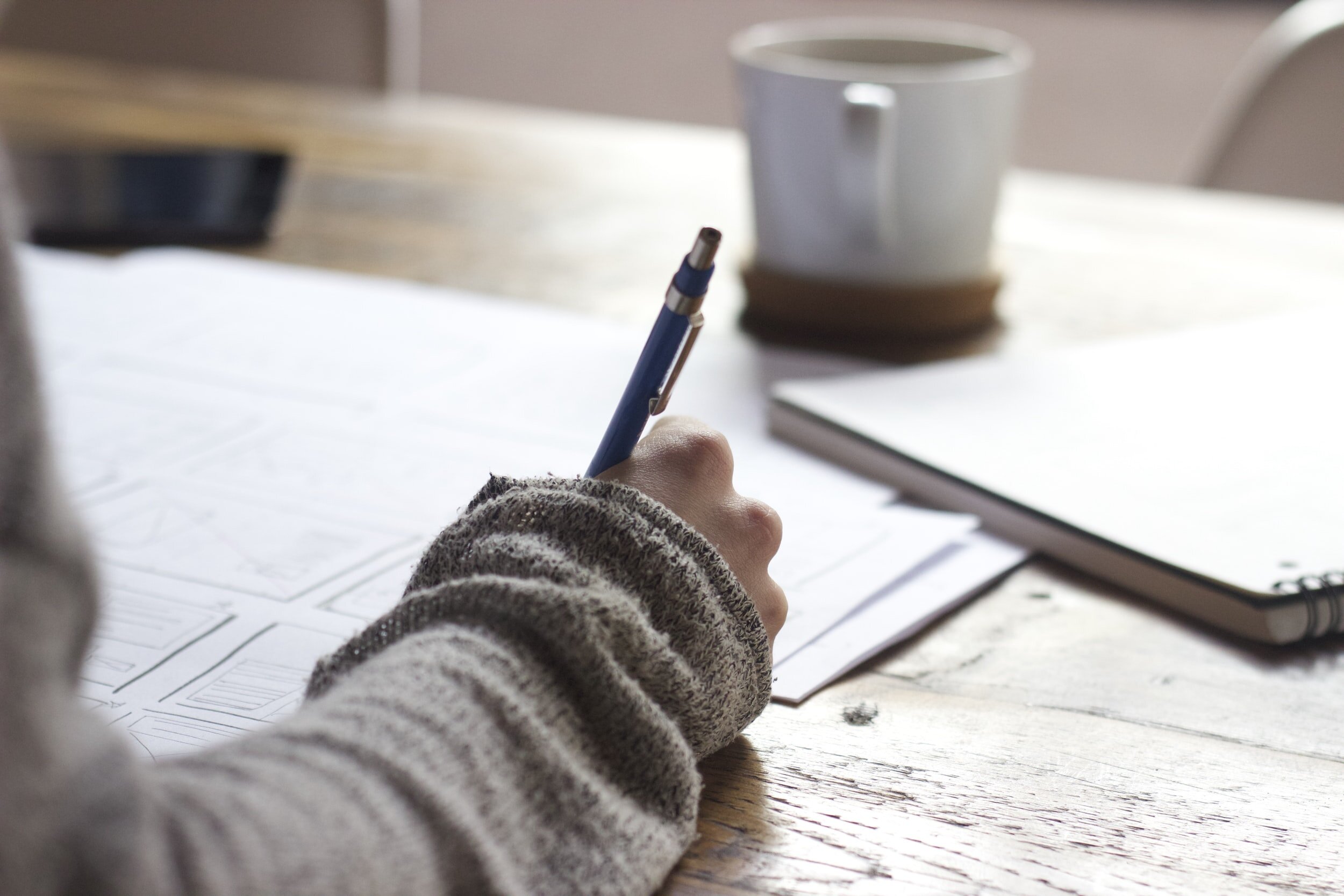 A hand writing with a pen at a desk beside a mug of coffee